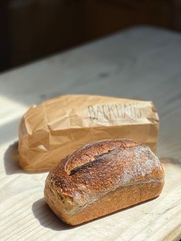 Close-up of a freshly baked artisan bread loaf next to a labeled paper bag, elegantly sitting on a wooden surface.