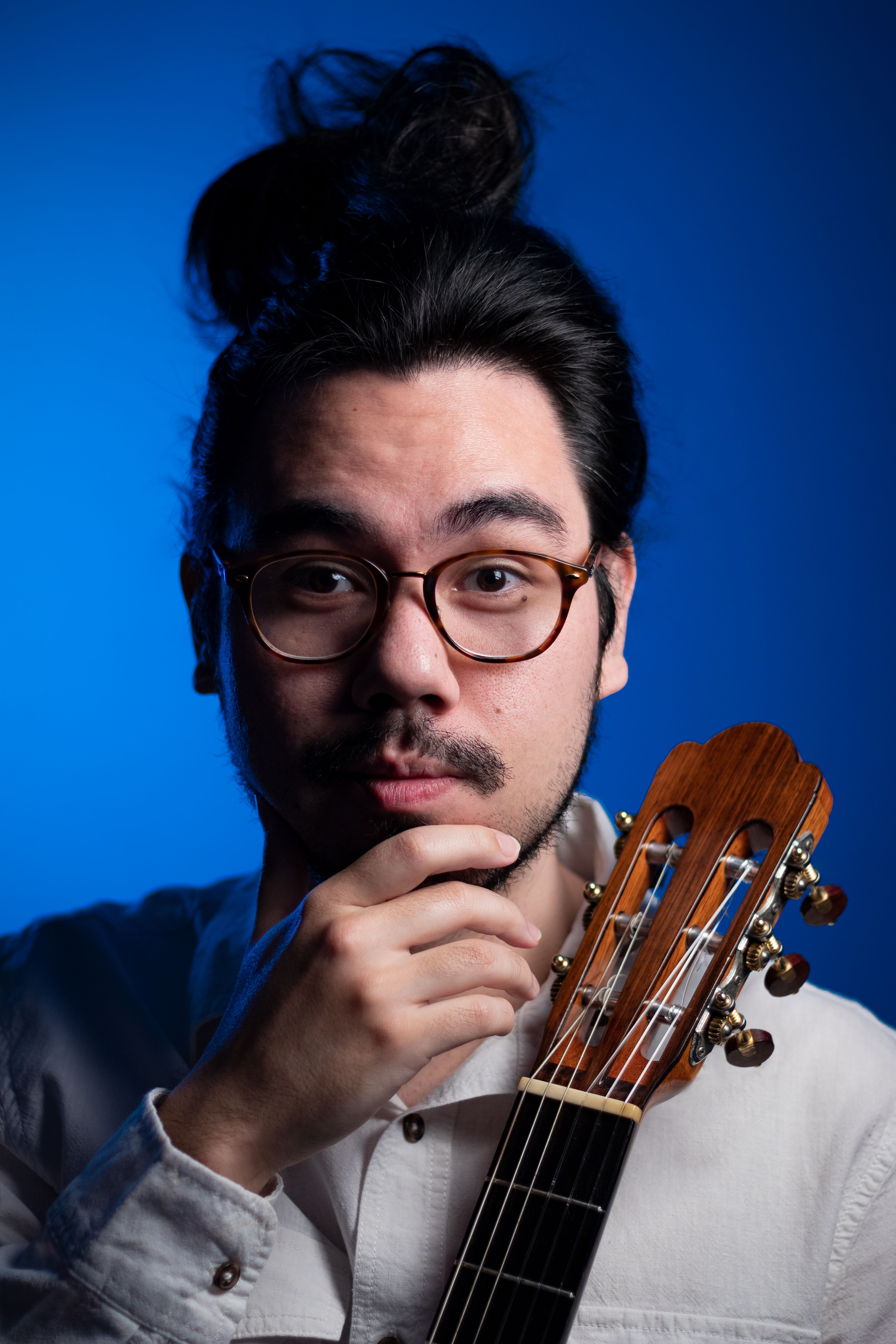 Studio portrait of a male musician with acoustic guitar, eyeglasses, and man bun.