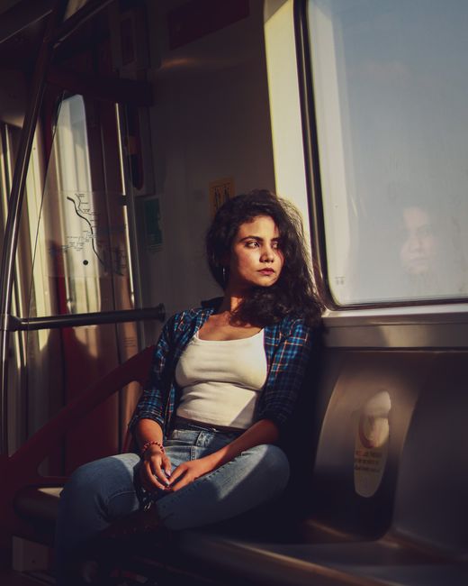 South Asian woman in plaid shirt sitting by window, staring outside during train journey in Ahmedabad.
