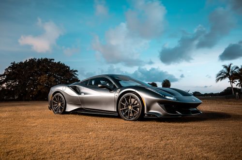 Free A sleek gray Ferrari parked on a grassy field under a bright blue sky with white clouds. Stock Photo