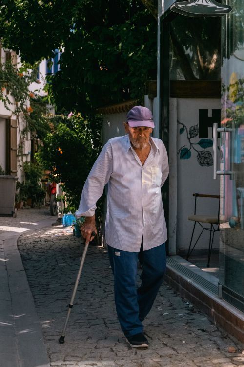 Free An elderly man wearing a cap and long sleeves walks with a cane down a sunlit alley. Stock Photo