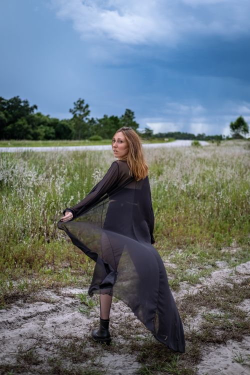 Free Elegant woman in a sheer black dress posing in a rural field under stormy skies. Stock Photo