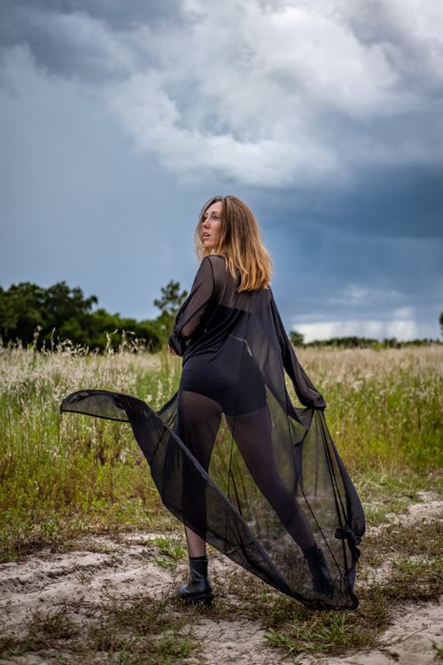Free A woman in a sheer black outfit poses in a scenic meadow under a dramatic cloudy sky. Stock Photo