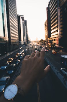 A hand with a wristwatch gestures towards a bustling city street during sunset.