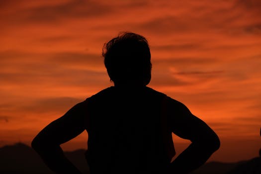 Silhouette of a person standing against a vibrant orange sunset, creating a dramatic sky scene.