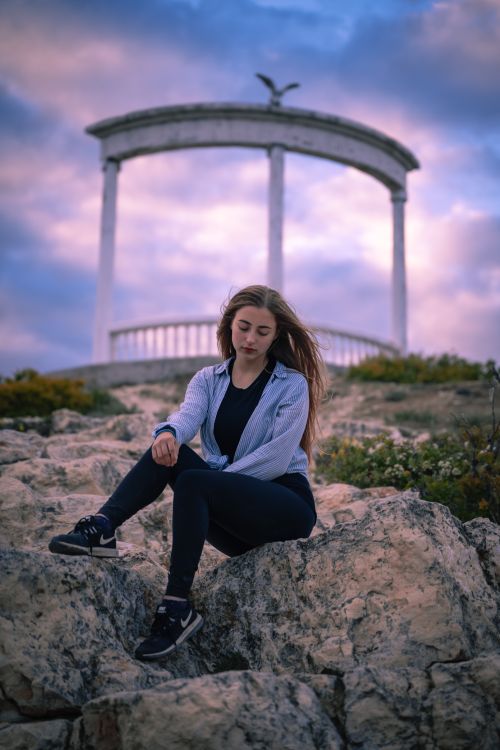 Free A young woman sitting peacefully on rocks with an architectural structure in the background during twilight. Stock Photo