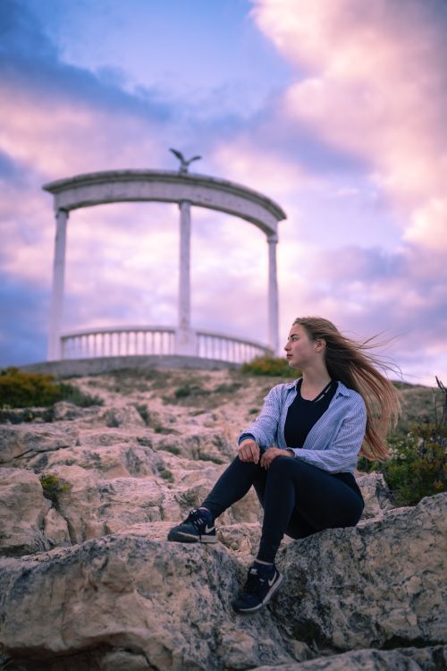 Free Woman sitting on rocks near an arch monument with a sunset backdrop. Stock Photo