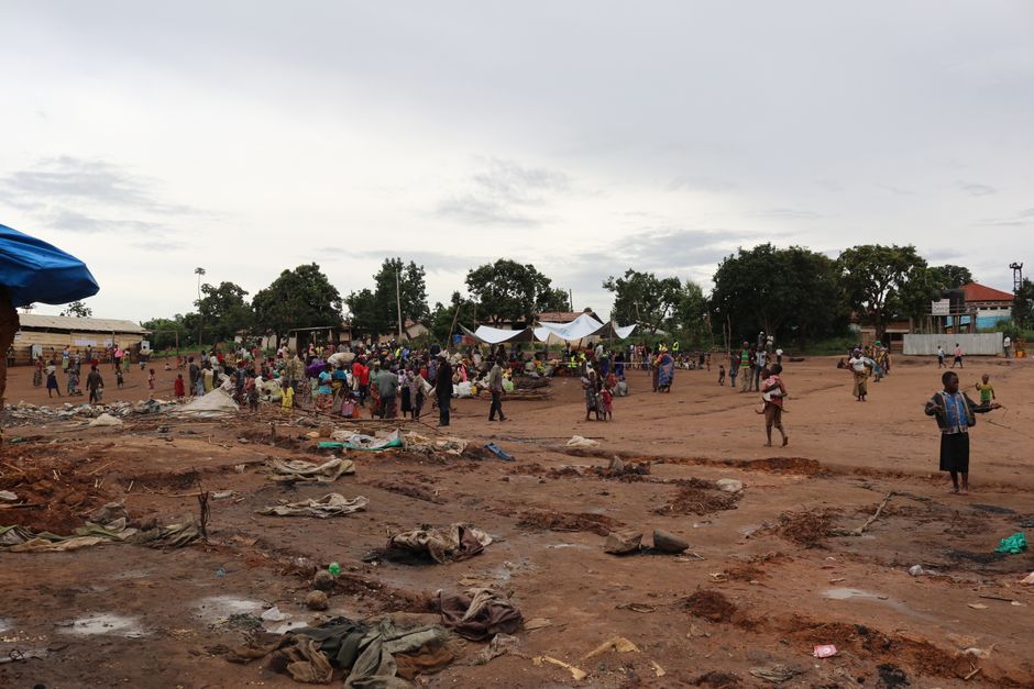 People gather in a large open area for a community event on a dirt ground.