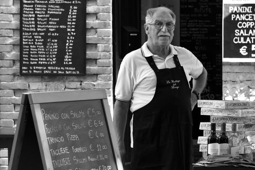 Senior man in apron by Grazzano Visconti food stall with menu boards.