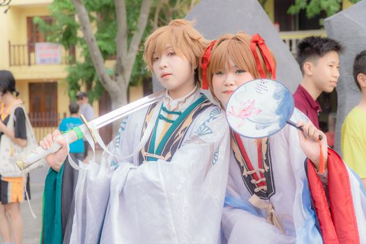 Two cosplayers in ornate fantasy costumes pose with swords and fans outdoors.