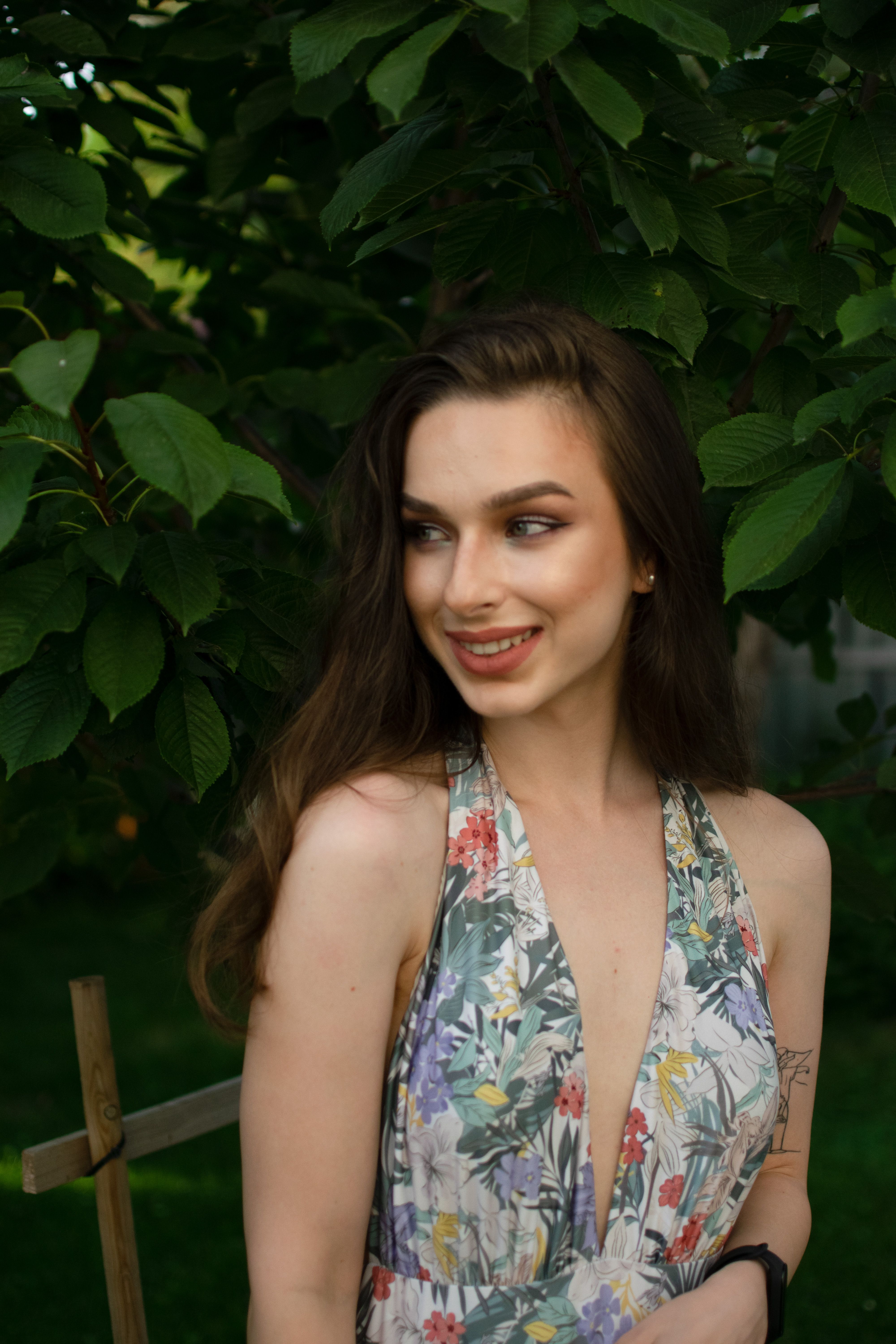 A smiling woman in a floral dress enjoying a serene outdoor setting under lush greenery.
