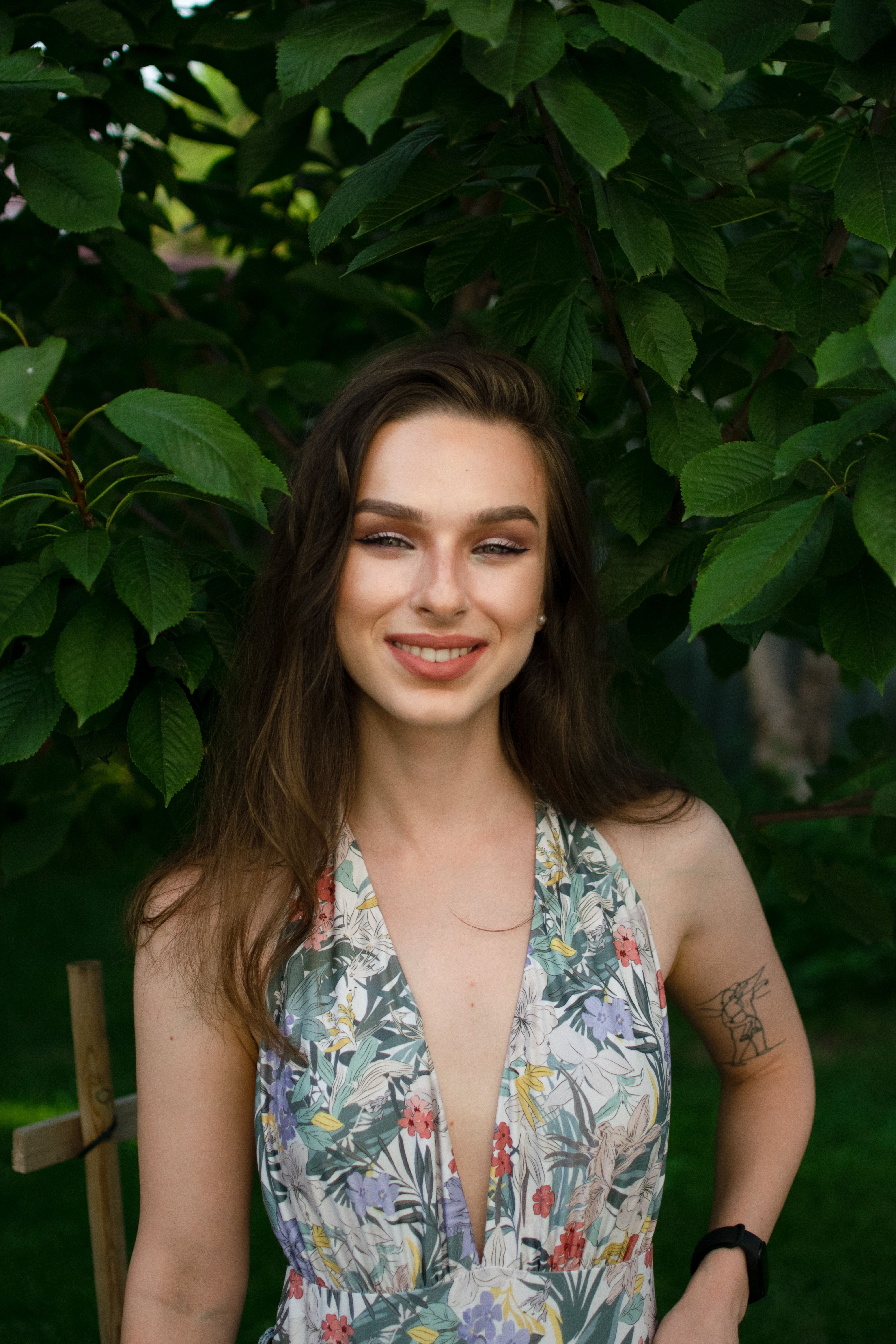 Portrait of a smiling woman in a floral dress standing outside in lush greenery.
