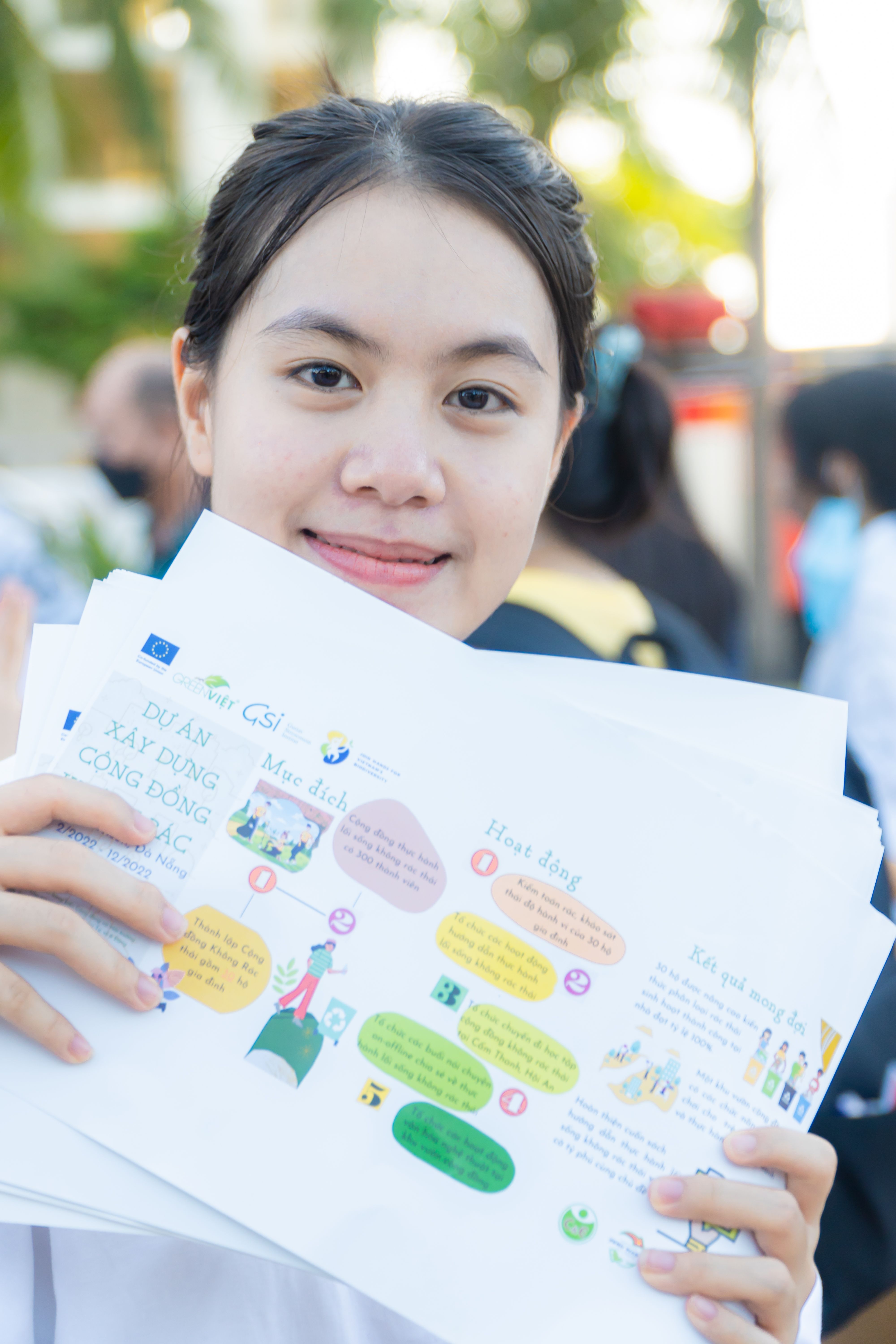 Smiling woman outdoors holding vibrant educational papers, in a lively setting.