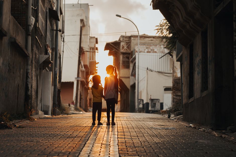 Dos niños caminando en un callejón iluminado por el sol en un entorno urbano al atardecer.