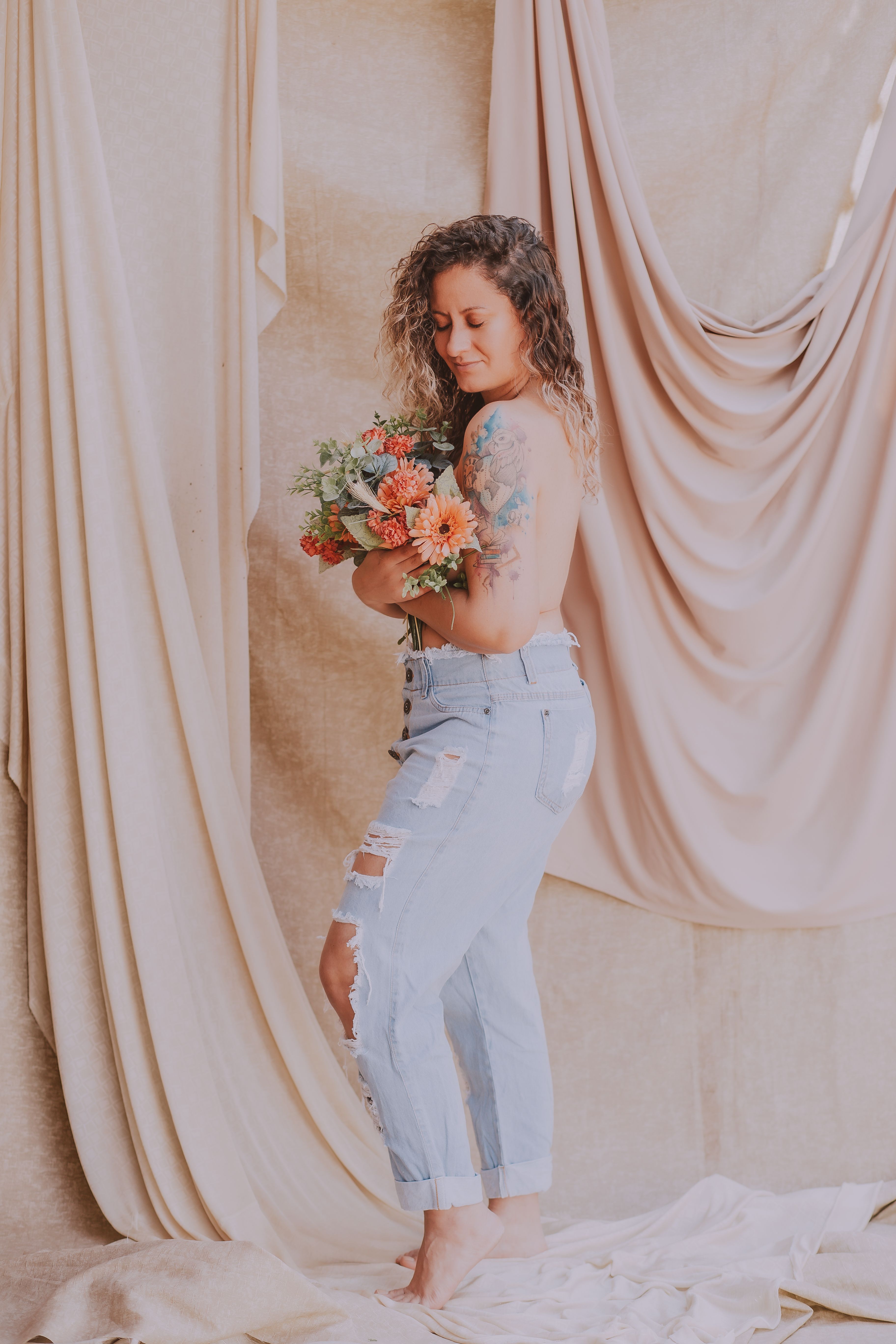 A tattooed woman poses topless with flowers in a soft, artistic setting.