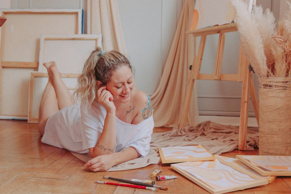 Como a Criatividade Pode Enriquecer sua Vida Pessoal e Profissional 2 Smiling woman relaxing and creating art in a cozy room with canvases and paints.