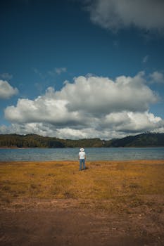 A person stands alone by a tranquil lake under a cloudy sky, evoking solitude and peace.