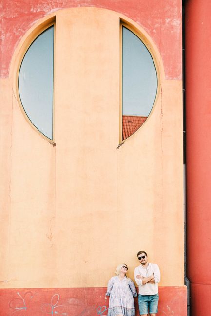 A couple stands casually against a vibrant architectural wall featuring unique circular windows.