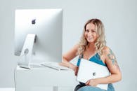 Smiling Woman Holding a Laptop Sitting Near a Desktop Computer