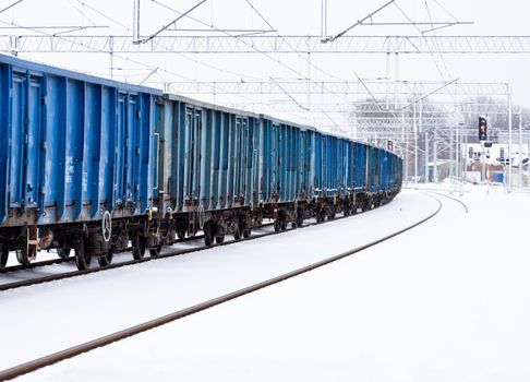 A blue cargo train travels on a snowy railway track in winter with overhead power lines.
