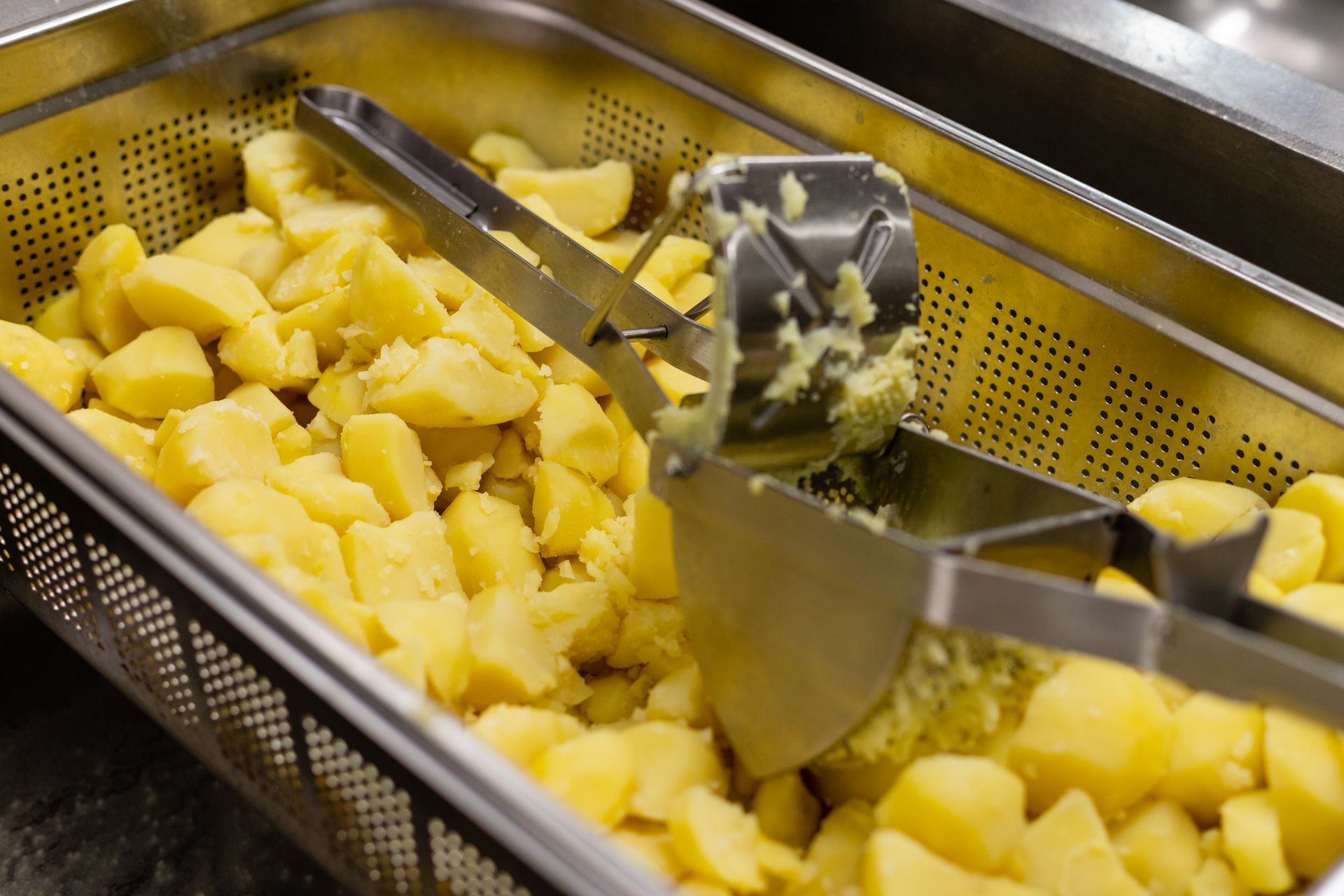 Steaming yellow potatoes being mashed with a metal potato ricer in a professional kitchen setting.