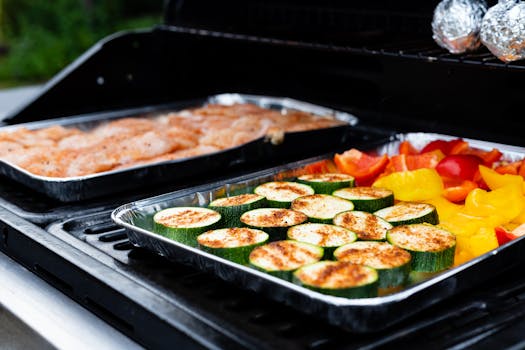 Colorful vegetables and seasoned chicken grilling on an outdoor barbecue in foil trays.