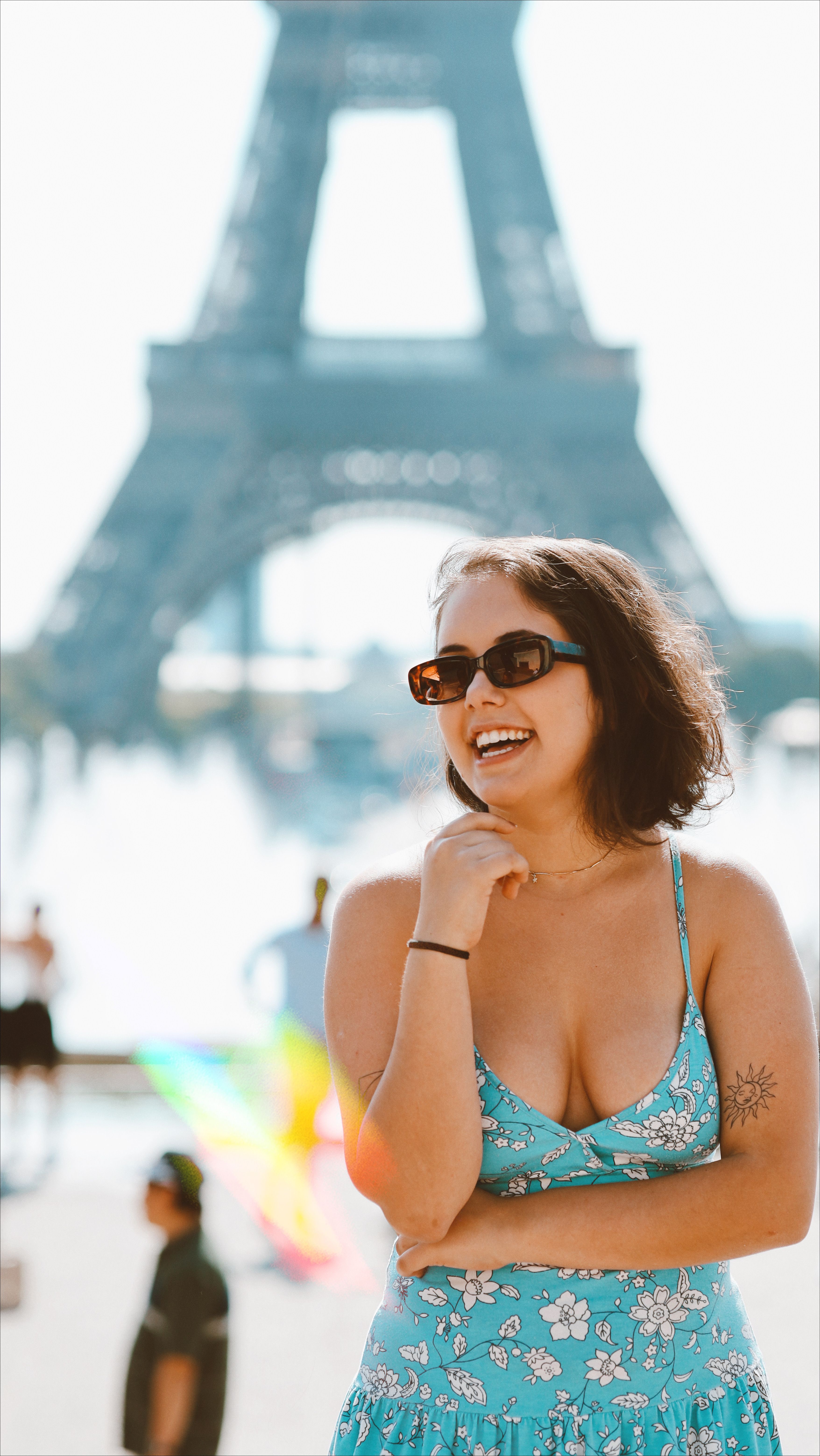Smiling woman in a floral dress poses in front of the Eiffel Tower on a sunny day.