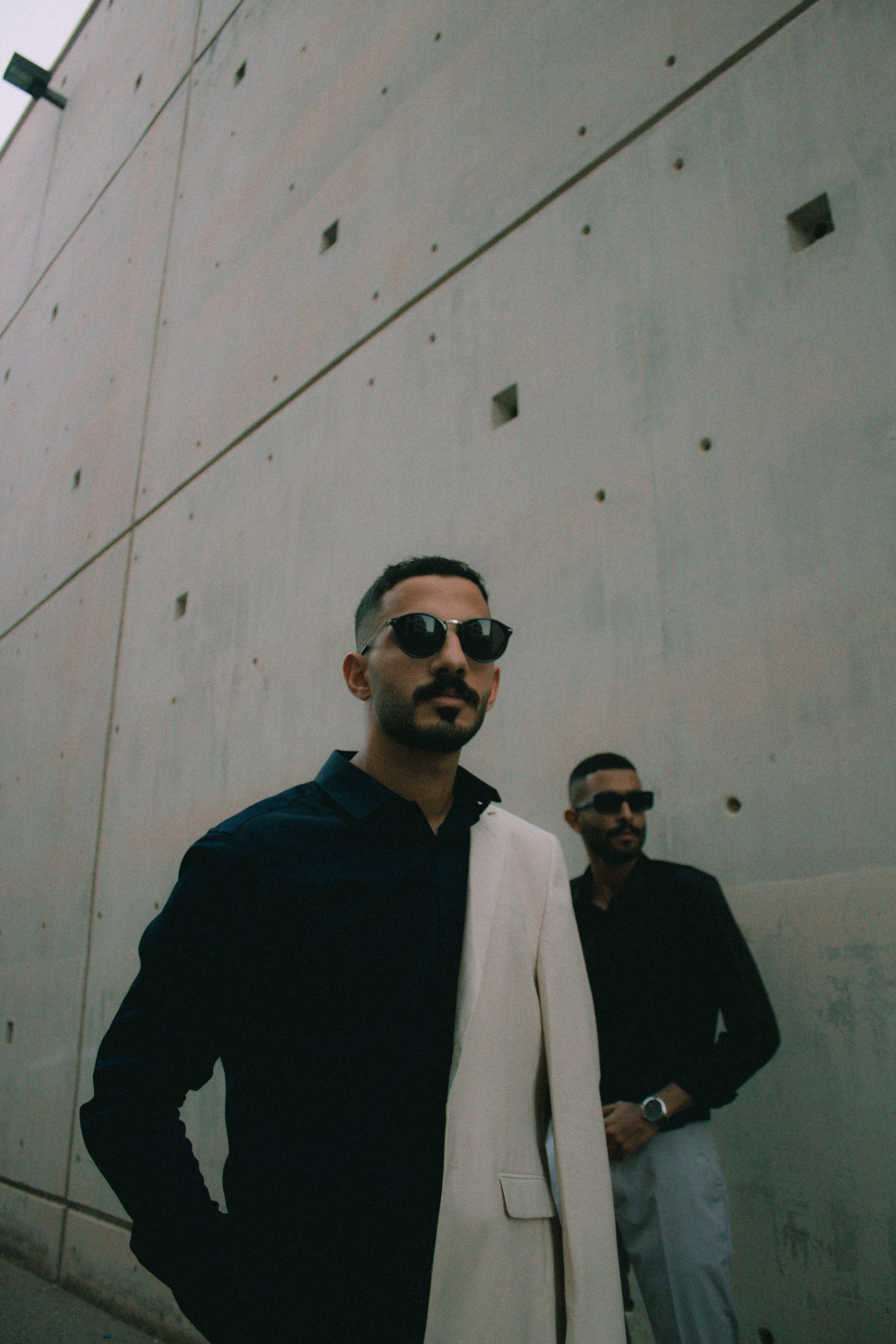Two fashionable men in sunglasses walking against a geometric wall in Muscat, Oman.