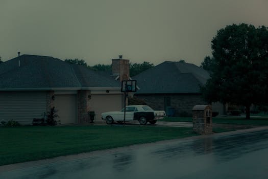 A classic car parked outside a suburban house near a basketball hoop on a wet road.