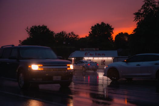 Vehicles driving on a wet road at sunset near a neon-lit inn, creating a moody travel scene.