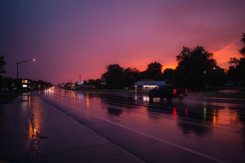 Free A wet road at twilight featuring a moving pickup truck and vibrant sky reflections. Stock Photo
