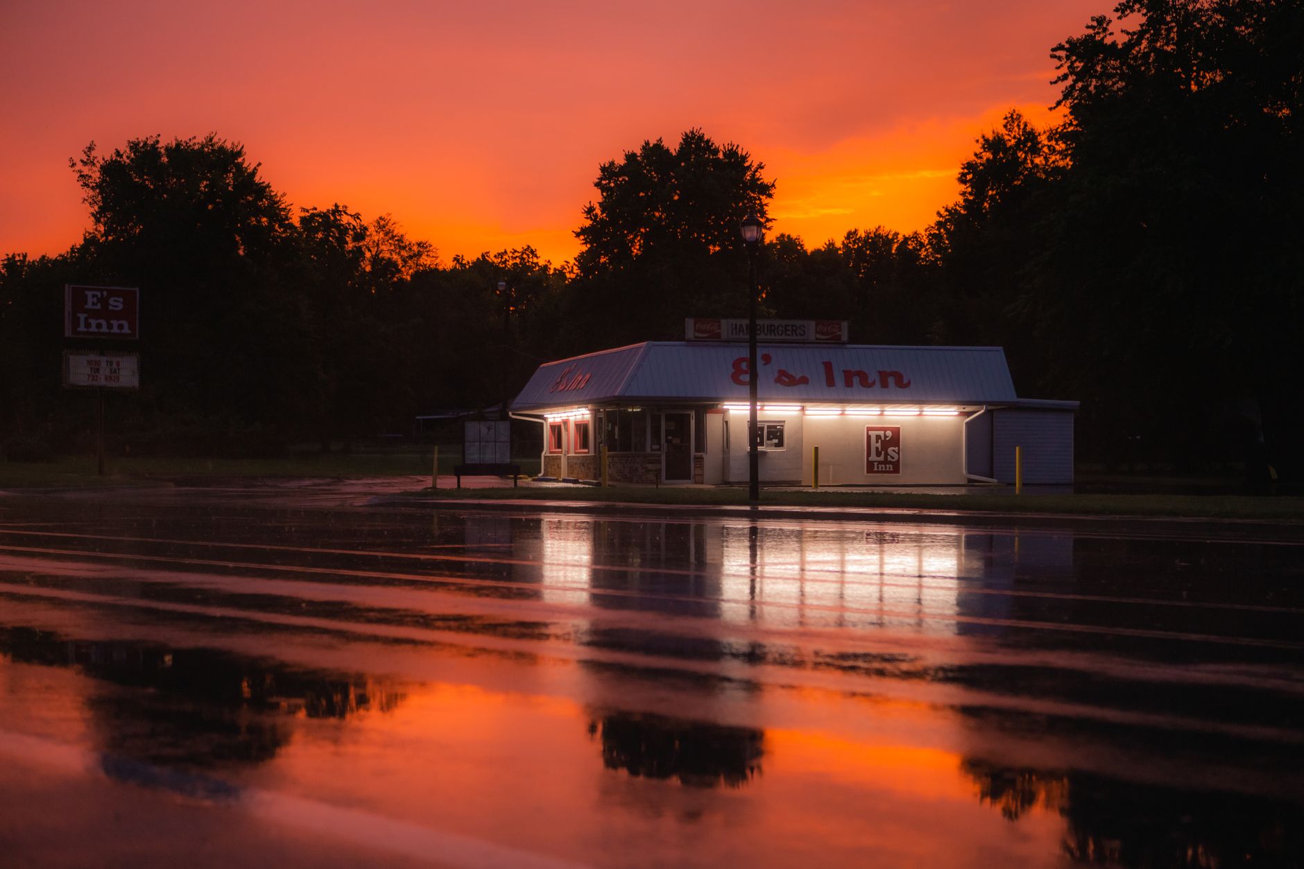 A roadside diner with neon signs reflecting on a wet street under a vibrant sunset sky.