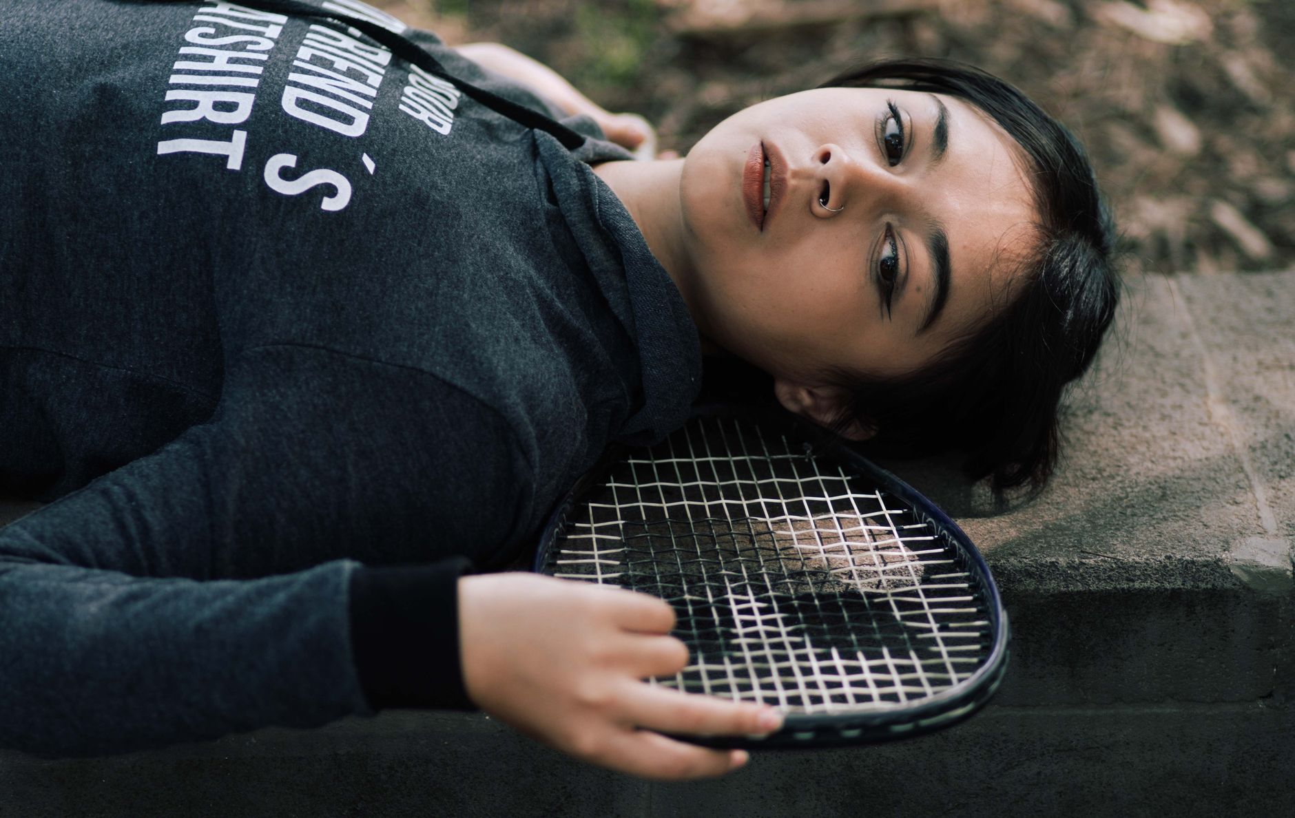 A woman in a black hoodie lies outdoors with a tennis racket, exuding a relaxed and pensive mood.