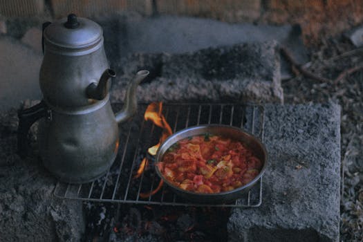 Rustic outdoor cooking setup with a vintage teapot and a skillet dish on a grill over an open flame.