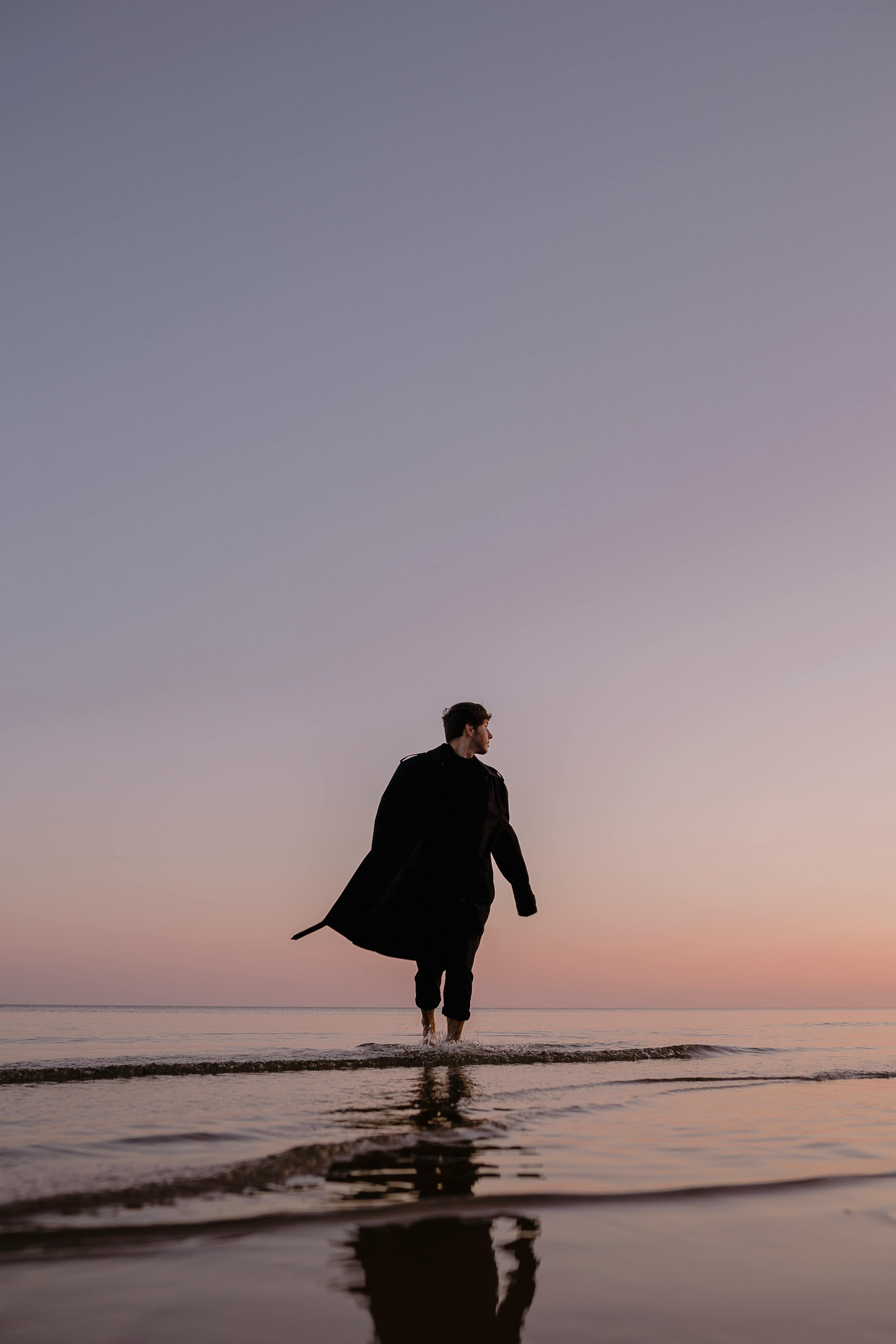 A man walking along the shore at twilight, captured with serene sea reflections.