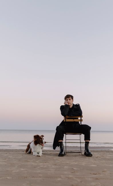 A young man sitting on a chair with a dog by his side on a tranquil beach at sunset.