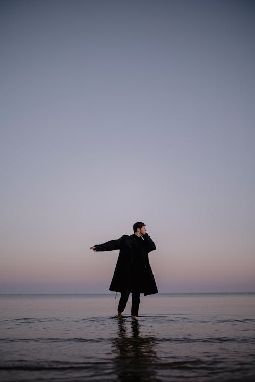 Man in Long Coat Standing in Water on the Beach 