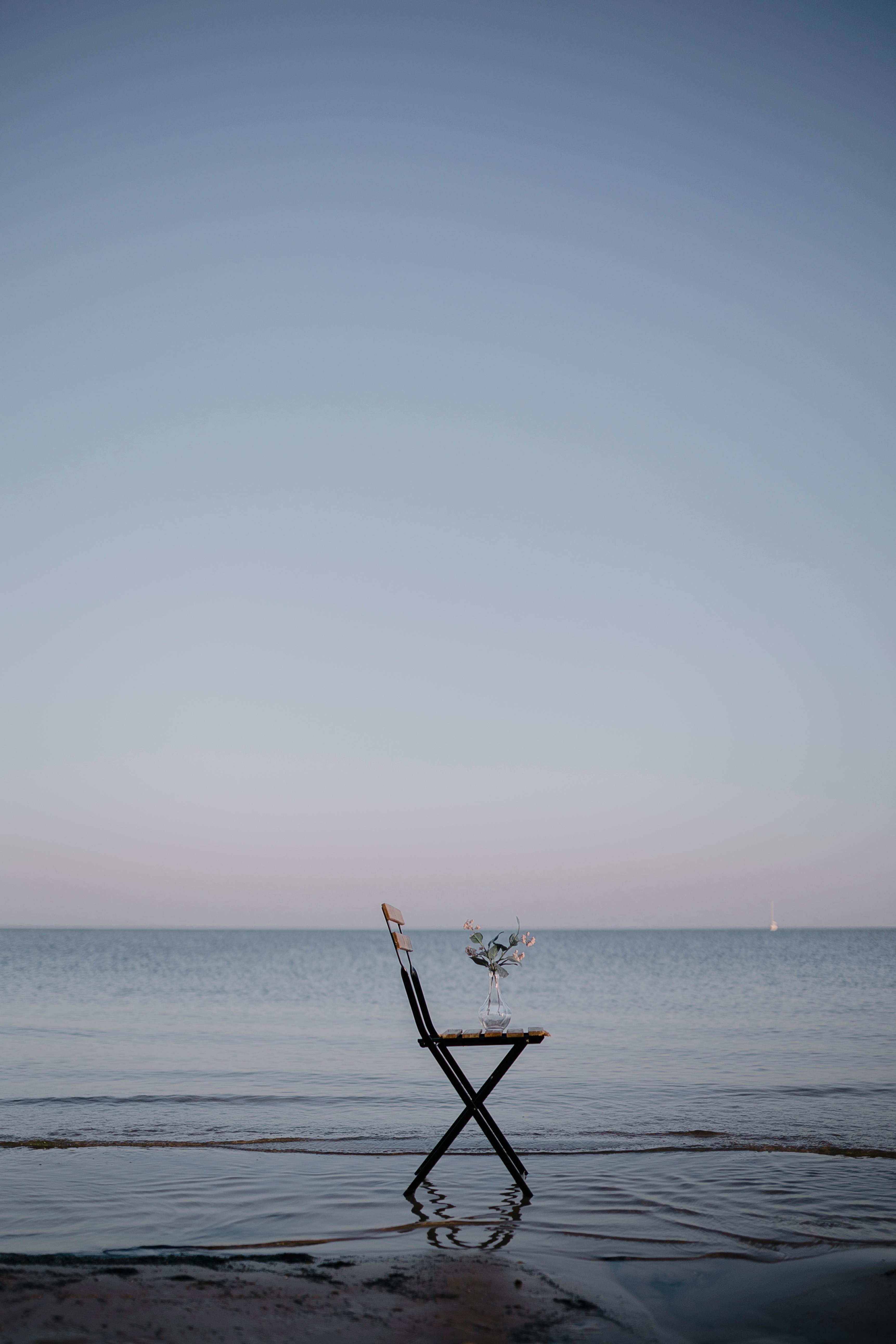 A wooden chair with a glass vase sits alone by the calm sea at dusk.