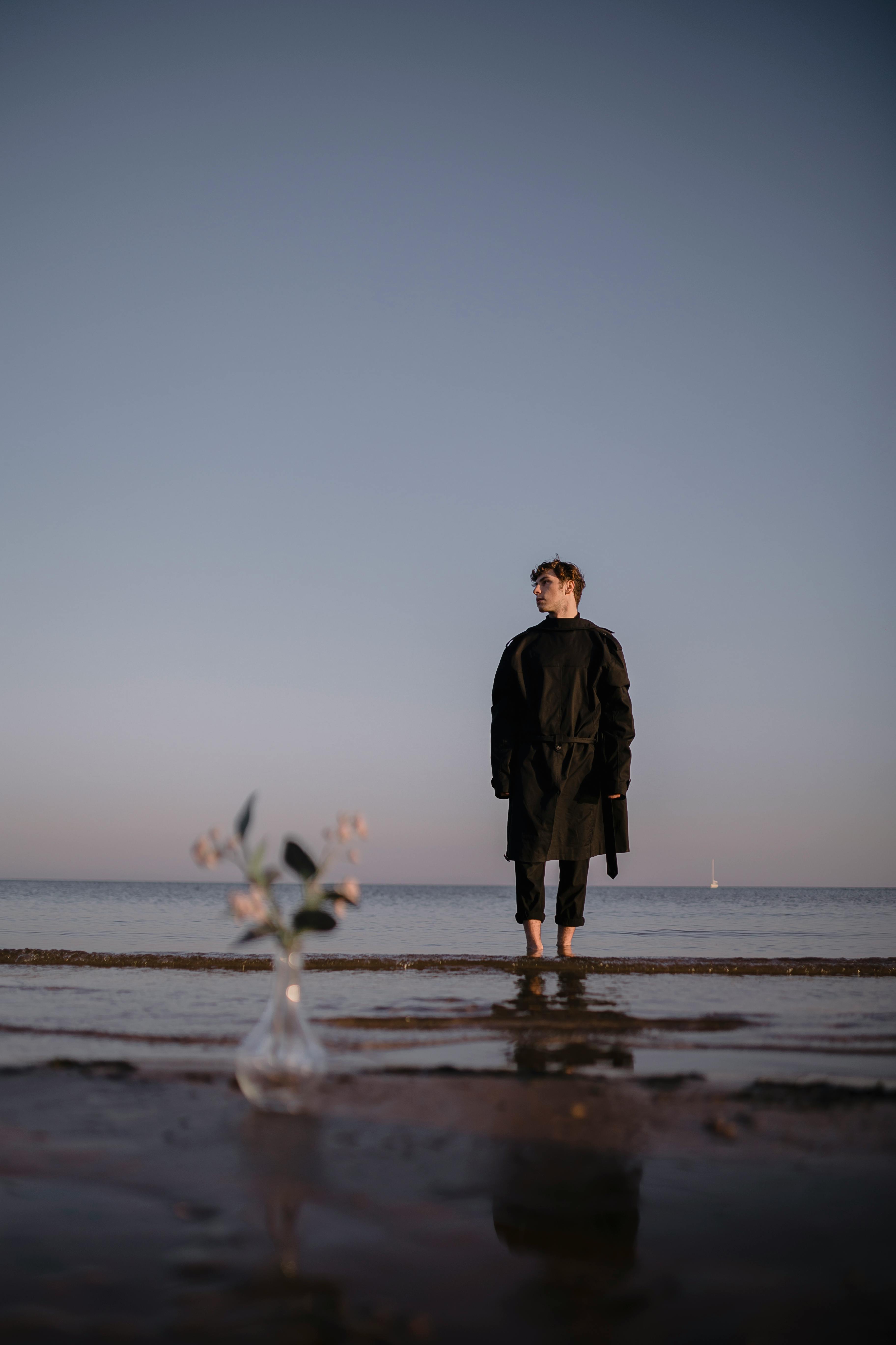 A man stands barefoot on a peaceful beach at dusk, with a vase of flowers in the foreground.