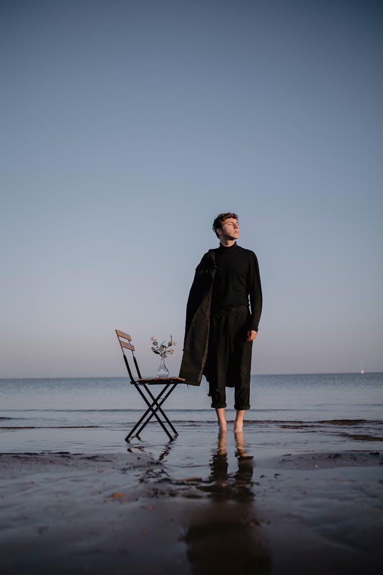 Man Standing On The Beach Next To A Chair With Flower Vase On It 