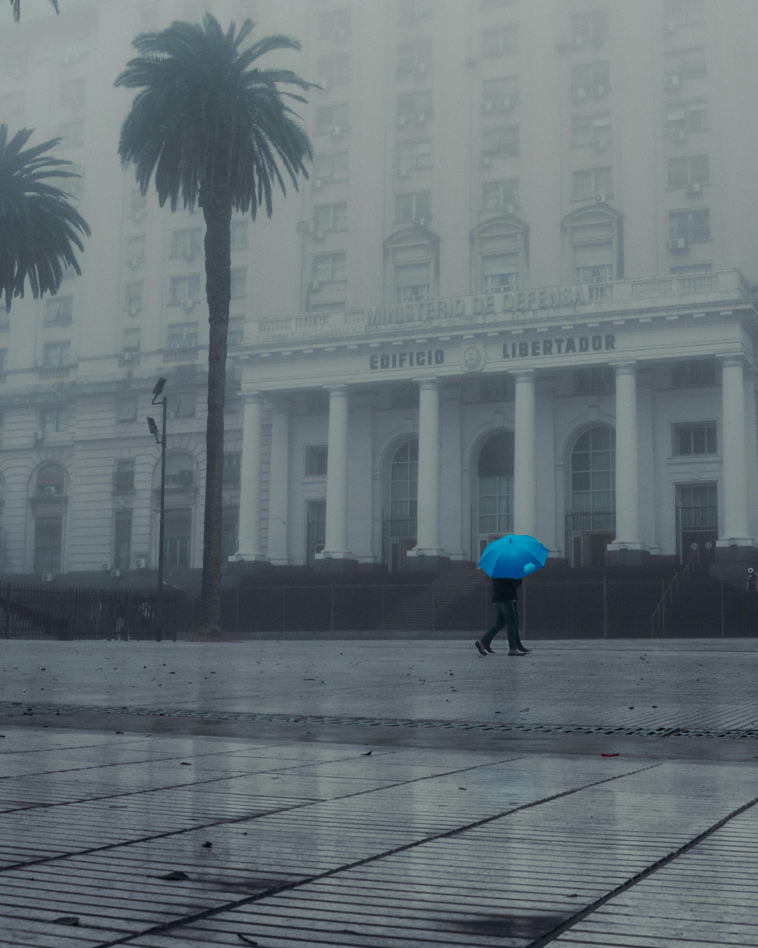 Man with blue umbrella walking past the foggy Edificio Libertador in Buenos Aires.