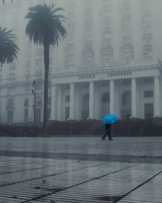 Man with blue umbrella walking past the foggy Edificio Libertador in Buenos Aires.