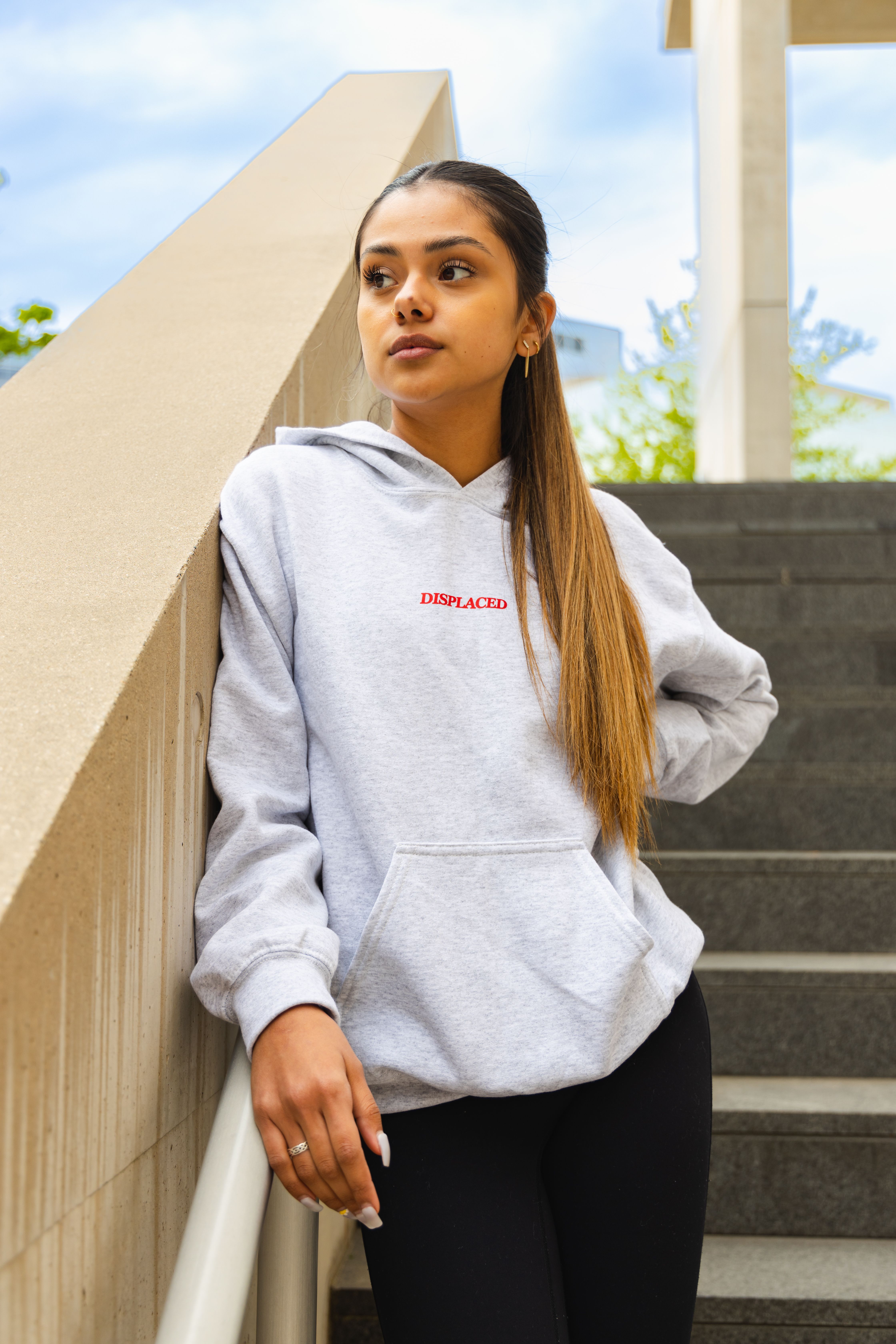 Young woman in gray hoodie stands outdoors by a staircase.