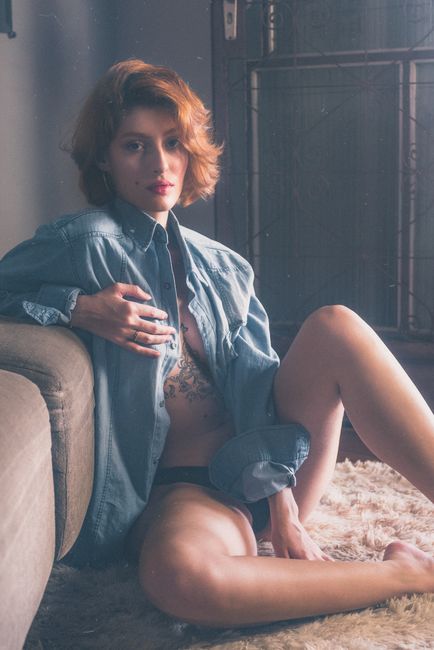 A woman with tattoos poses indoors in a denim jacket and black bikini, seated on a fur carpet.