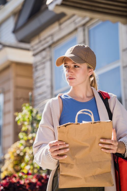 Free A young woman stands outdoors holding a paper bag, looking away thoughtfully in a residential street. Stock Photo