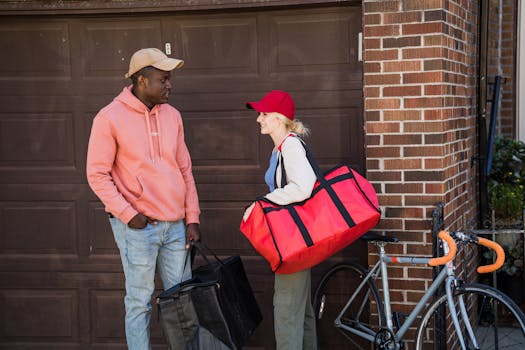 Two delivery workers chat outside a garage, each carrying bags and a nearby bicycle.