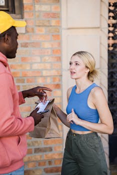 A delivery man hands a paper bag to a woman outside a building.