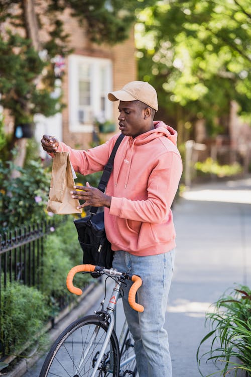 Delivery Man on a Bicycle Holding a Paper Back with an Order and Standing on a Sidewalk in Front of a House 