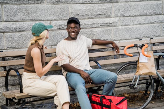 A man and woman laughing while sitting on a bench next to a bicycle and delivery bag.