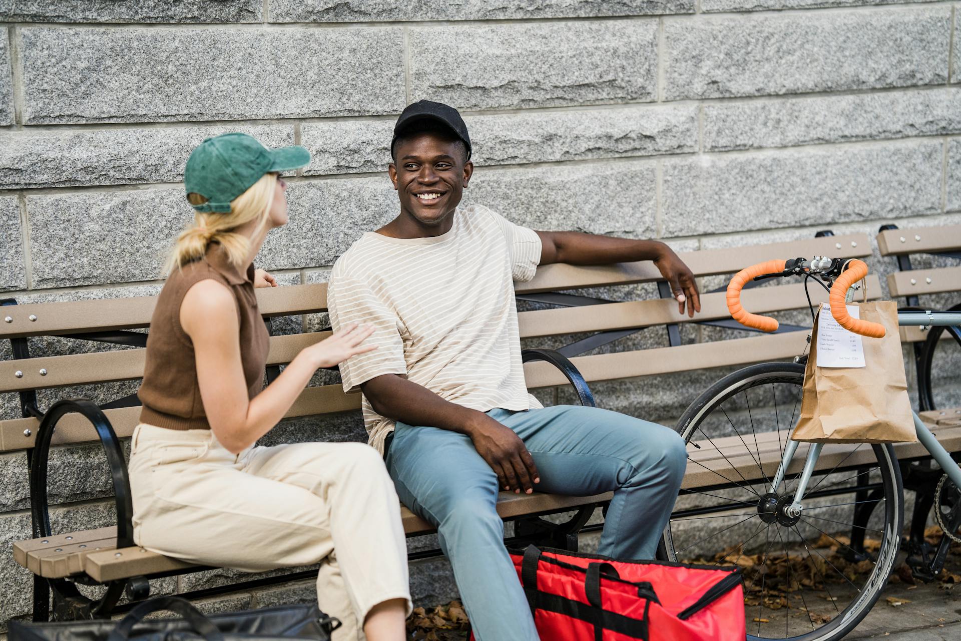 Young Man and Woman Working in Food Delivery Services Sitting on a Bench in City Talking and Smiling